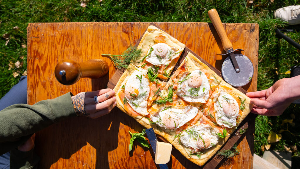 Two hands reaching to grab a piece of breakfast pizza on a picnic table with grass in the background. The vibrant outdoor scene highlights the delicious pizza, perfect for a picnic setting. 