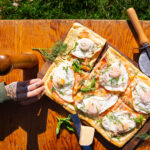 Two hands reaching to grab a piece of breakfast pizza on a picnic table with grass in the background. The vibrant outdoor scene highlights the delicious pizza, perfect for a picnic setting.