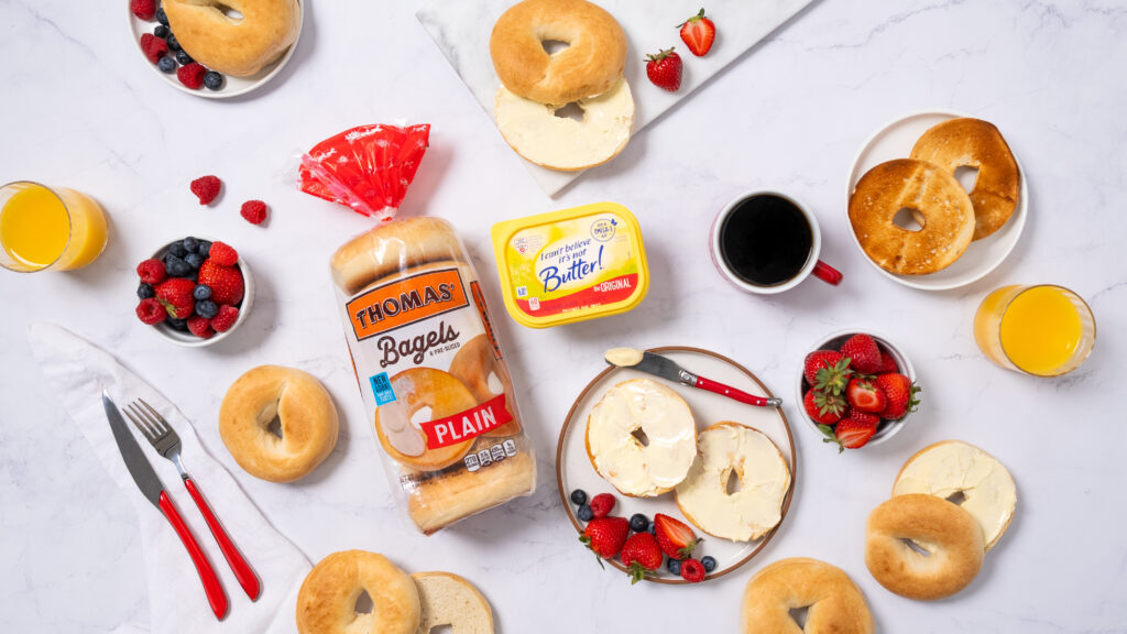 Breakfast scene featuring bagels with butter spread, fresh fruit, and coffee. CPG food brand product featured in center of frame with berries and silverware to accent. 