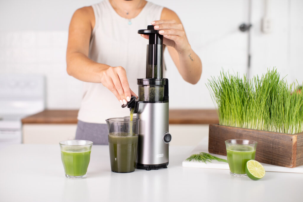 Brand marketing photo of a studio kitchen scene featuring a hand model using a juicer with green juice on display. Wheatgrass styled around the setup enhances the fresh and healthy vibe. Ideal for showcasing nutritious beverage preparation through professional food videography and photography, highlighting the appeal of your health products.