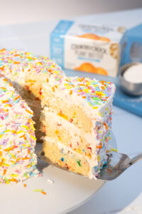 Lovely layered cake slice with sprinkles being pulled out to be plated with featured product in the background.