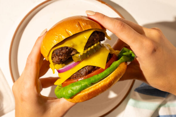 Burger with Cheddar Close-up of a gourmet burger featuring fresh lettuce, cheese, tomato, and onion, held in two hands to emphasize its juicy and appetizing ingredients. Perfectly captured to showcase the quality and appeal of the product featured.