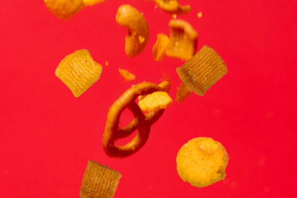 Crunch Mix Falling Clear photograph of snack mix captured mid-air against a red backdrop, highlighting the texture and visual appeal of the product.