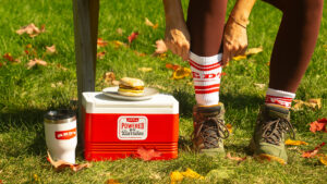 A sandwich (the product) featured atop a cooler in an outdoor fall setting, with a model sitting on a bench nearby, ready to embark on a hike, showcasing the product's convenience.