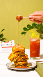 Food photography image of a stack of breakfast sandwiches drizzled with honey, accompanied by a Bloody Mary in the background, on a green backdrop.