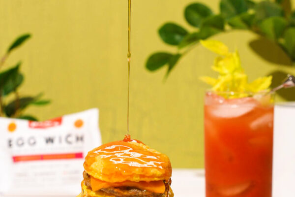 Hot Honey Drizzle Over Breakfast Sandwiches Stack of breakfast sandwiches drizzled with honey, accompanied by a bloody mary in the background, on a green surface.