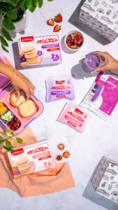 Marble surface with pink accents highlighting the product, styled with lunch boxes, smoothies, and hand models on each side.