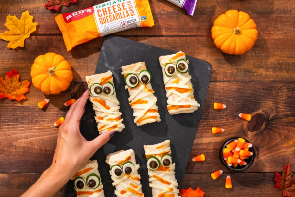 Halloween Burritos Halloween-themed burritos styled as mummies with seasonal fall leaves and pumpkins, featuring a hand model reaching in, all on a wooden surface.