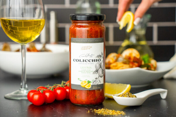 Clam Ragout with Pancetta Pasta sauce featured in front of a pasta dish with a glass of wine. On a studio set with black brick and black marble.