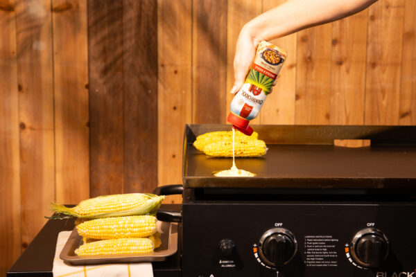 Grilling Butter Spread Product being poured onto a blacktop grill with a wooden fence in the background. A hand model is shown pouring the product, alongside featured corn, capturing the essence of outdoor grilling.