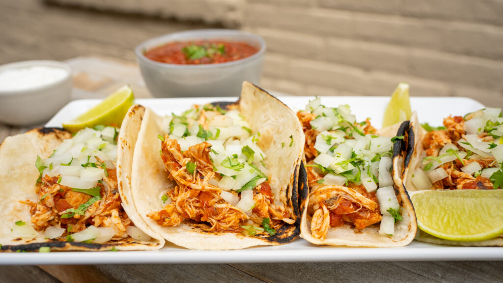 Food photography of chicken tacos topped with onion and cilantro with salsa int the background. Photo taken outside to utilize natural lighting. 