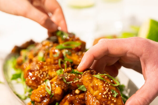 Miso Chicken Wings Hands reaching for sauced chicken wings on a white background, showcasing the delicious and shareable appeal of the product.