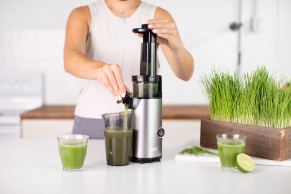 Studio-lit kitchen scene featuring a hand model using a juicer, showcasing green juice with wheatgrass accents, highlighting the product's fresh and healthy appeal for food brand marketing.